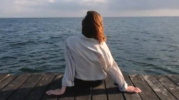 Dziewczyna lub młoda kobieta siedzi na drewnianych molo i patrzy na morze. Koszula na dziewczynie i jej włosy trzepotające na wietrze — Wideo stockowe