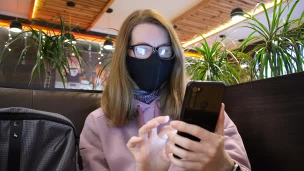 Gadis atau wanita muda bekerja pada ponsel di kafe. Wanita bertopeng pelindung di wajahnya di kantin berguling-guling melalui jaringan sosial — Stok Video