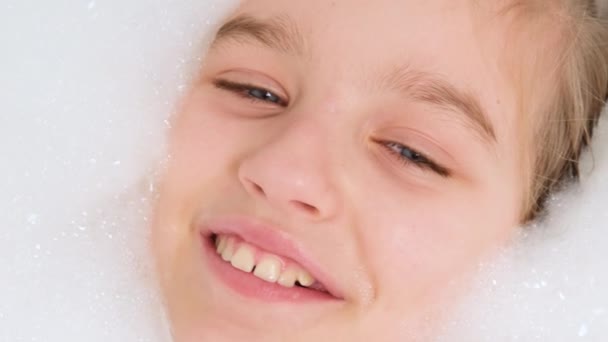 Портрет веселой девочки-подростка, лежащей в ванне с густой мыльной пеной и смотрящей в камеру. Концепция детской гигиены и здравоохранения на дому. Ребенок развлекается и играет дома — стоковое видео