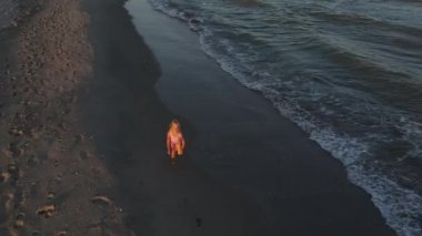 9 yaşında kırmızı elbiseli bir kız ıssız bir sahilde su hattı boyunca koşuyor. İHA saldırısı. Üst Manzara. Çocuk koş ve deniz kıyısında oyna.