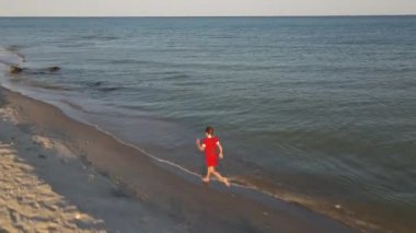 9 yaşında kırmızı elbiseli bir kız ıssız bir sahilde su hattı boyunca koşuyor. İHA saldırısı. Üst Manzara. Çocuk koş ve deniz kıyısında oyna.