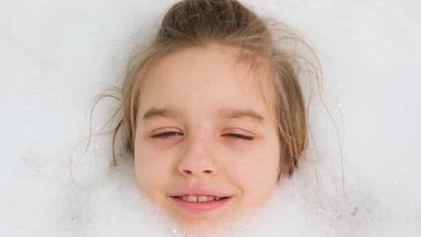 Kalın sabun köpüğüyle banyoda yatıp kameraya bakan neşeli genç kızın portresi. Evde çocuk hijyeni ve sağlık hizmetleri kavramı. Çocuk eğleniyor ve evde oynuyor. — Stok video