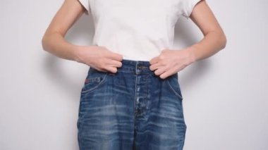 Beyaz arka planda büyük beden kot pantolonla dans eden mutlu kadın. Diyet konsepti, düzgün beslenme, kilo kaybı ve vücut pozitifliği. Sıska Kadın Gevşek Kotları ve Zayıflığını Gösteriyor