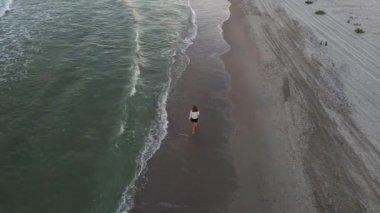 Güzel çıplak ayaklı kadın ıssız sahilde yürüyor. Deniz sahilinde, sahil şeridinde ve yürüyen kızda insansız hava aracı görüntüsü. 4K çözünürlük videosu.