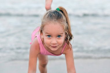 Little happy and smiling girl performs gymnastic and dance exercises and poses against the backdrop of the sea and beach clipart