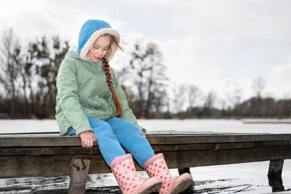 Ragazza sognante seduta sul ponte e immergere stivali di gomma nell'acqua del fiume. Bandiera di inizio primavera con spazio di copia — Foto Stock