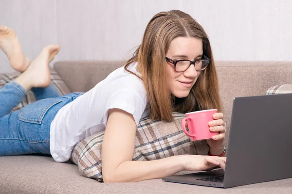 젊은 프리랜서 여성 이 노트북으로 일하고 있습니다. 여성은 비디오 콘퍼런스를 통해 가족 과 소통 한다. 차를 마시는 소녀 노트북에서 영화를 보고 있는 모습 — 스톡 사진