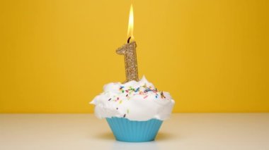 Birthday cupcake With Burning golden Candle number one on bright yellow Background. Slow motion first anniversary video banner.