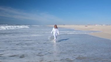 Genç bir kadın Atlantik Okyanusu boyunca koşuyor, eğleniyor ve gülüyor. Issız bir sahilde okyanus dalgalarının tadını çıkaran bir kız. Tam HD yavaş çekim videosu.