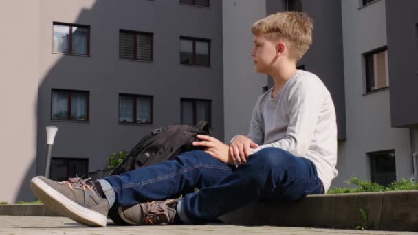 Schoolboy está sentado no passeio e olha em torno de um dia quente ensolarado — Vídeo de Stock