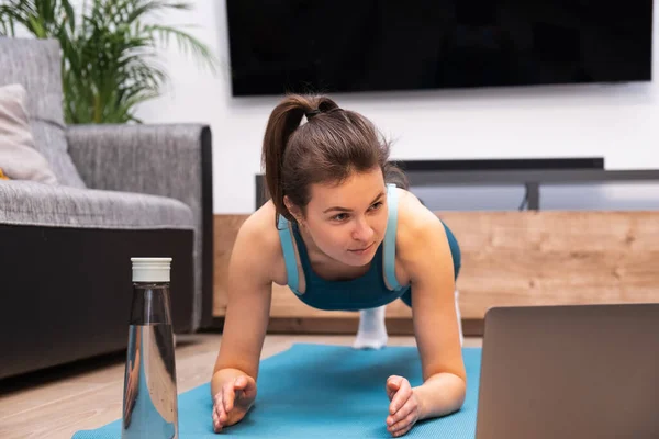 Woman trains at home, does the plank and watches the video on laptop at home — Stock Photo, Image