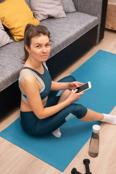 Ung kvinna i sportkläder tittar på kameran medan du sitter på en matta och håller i en smartphone — Stockfoto