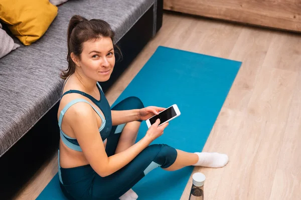 Усміхнена спортивна молода жінка використовує смартфон, сидячи на спортивному килимку вдома — стокове фото