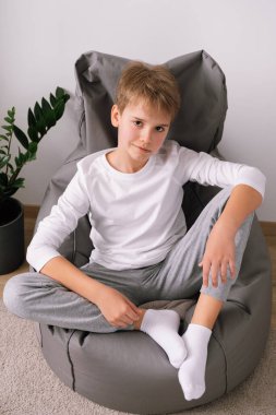 Sarışın, rahat stil sahibi bir genç, evdeki koltukta oturuyor. Beyaz tişört modeli.