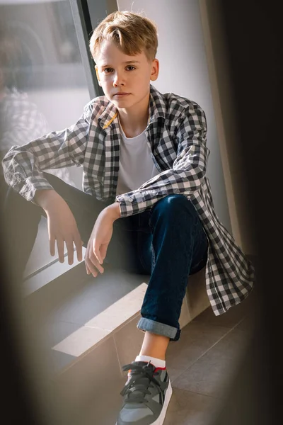 Stylový teenager v neformálním oblečení se dívá do kamery, zatímco sedí na podlaze v budově poblíž okna — Stock fotografie