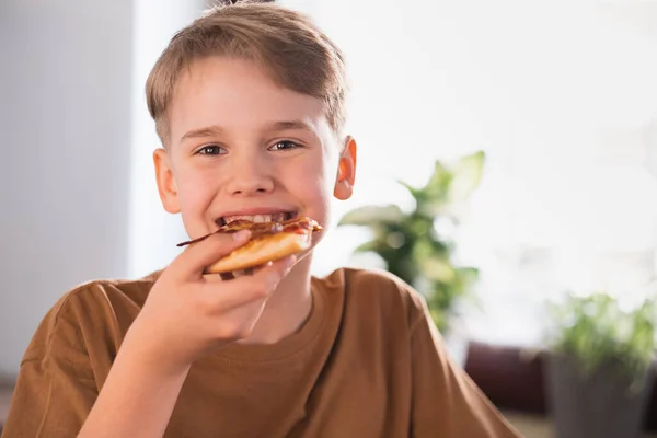 Zadowolony chłopiec je pyszną pizzę w kuchni w domu. — Zdjęcie stockowe