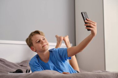 Rahatlamış mutlu çocuk yatakta yatarken selfie mi çekiyor yoksa video mu çekiyor? Şirin, rahat, mutlu çocuk portresi, yeni nesil taşınabilir hayat..