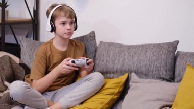 Pre-teen boy playing video game with joystick sitting on sofa at home.