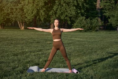 Genç bir kadın şehir parkında savaşçı pozu verirken yoga yapıyor. Açık hava sporları. Sağlıklı yaşam tarzı ve uzatma kavramı.