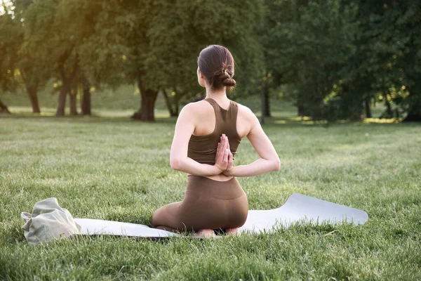 A young positive woman in a gymnastic suit practices yoga and meditates while sitting on a mat. Rear view. The concept of a healthy lifestyle and life extension