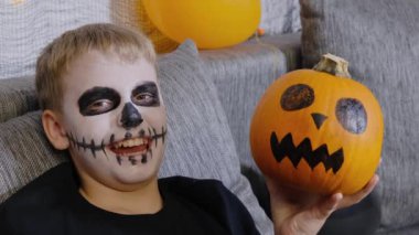 İskelet şeklinde makyajı olan ve elinde balkabağı olan korkunç bir çocuk kameraya bakıp gülüyor. Çocuk Cadılar Bayramı 'nı kutluyor.