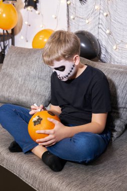 İskelet makyajlı bir çocuk evde kanepede otururken Cadılar Bayramı için balkabağı boyuyor. Çocuk şeker mi şaka mı tatiline hazır..