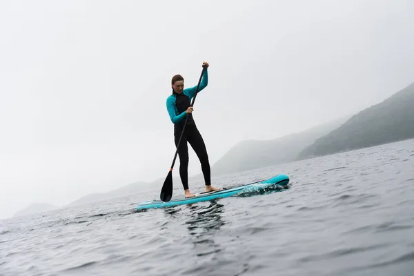 Mulher fitness em pé na prancha de surf desfrutando de supsurf com remo em névoa de água do mar sem fim — Fotografia de Stock