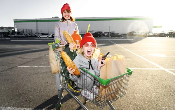 Mãe rola um menino e uma menina no carrinho de supermercado das crianças perto do supermercado — Fotografia de Stock