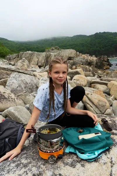 Весела дівчина-підліток-хіпстер, насолоджуючись табором, зупиняється на кам'яному пляжі, розслабляє приготування їжі на газі — стокове фото