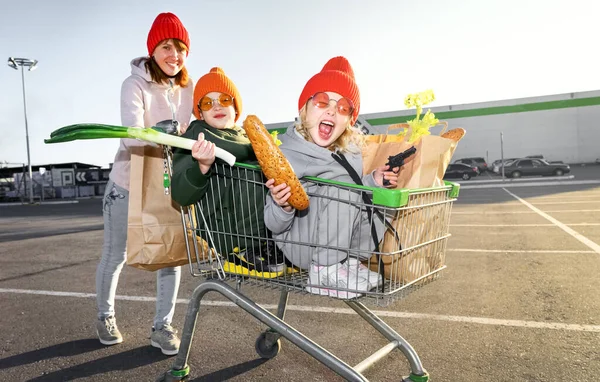 Mamá enrolla a un niño y una niña en el carrito de la compra de los niños cerca del supermercado — Foto de Stock