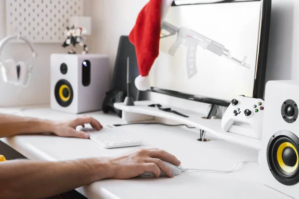男性手放在电脑桌上握着游戏鼠标和键盘。显示器上挂着一顶红色的圣诞老人帽. — 图库照片