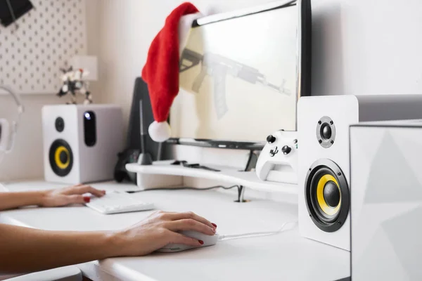 Женские руки на компьютерном столе держат игровую мышь и клавиатуру. Красная шляпа Санты висит на мониторе. — стоковое фото
