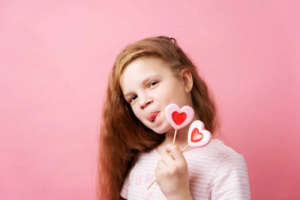 Menina segurando grandes pirulitos em forma de coração em suas mãos em um fundo rosa. — Fotografia de Stock