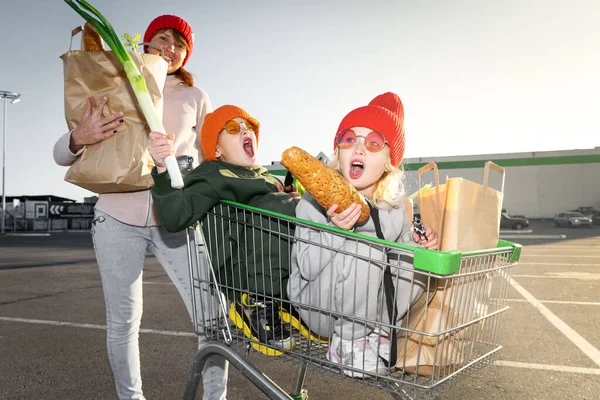 Mamá enrolla a un niño y una niña en el carrito de la compra de los niños cerca del supermercado — Foto de Stock