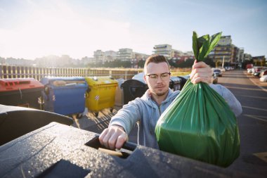 Çöplerle yürüyen bir adam. Şehrin caddesindeki çöp kutusuna plastik poşet atan kişinin ön görüntüsü..