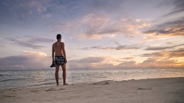 Cennet gibi beyaz kumsaldaki genç adamın arka görüntüsü. Mayolu turist Maldivler 'de renkli gün batımını izliyor