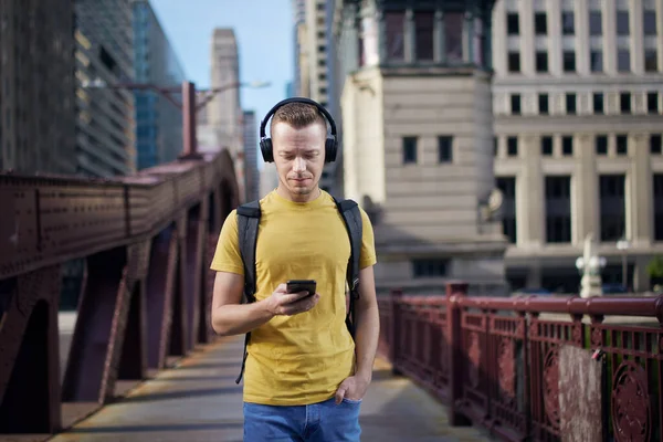 Young man listening to music with wireless headphones and looking into his cell phone during city walk.