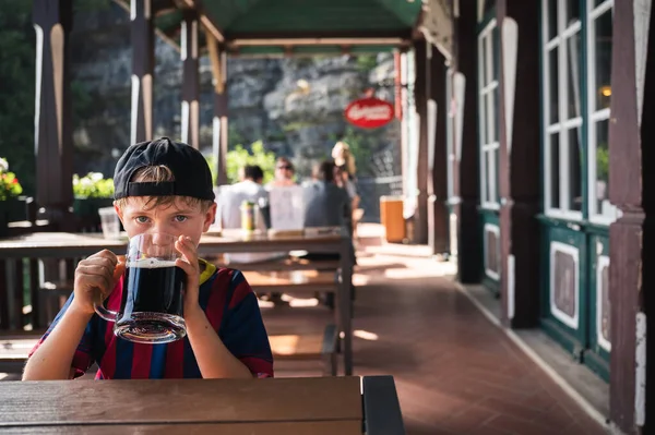 Cute boy is drinking a black drink from a large mug at a restaurant outdoor table
