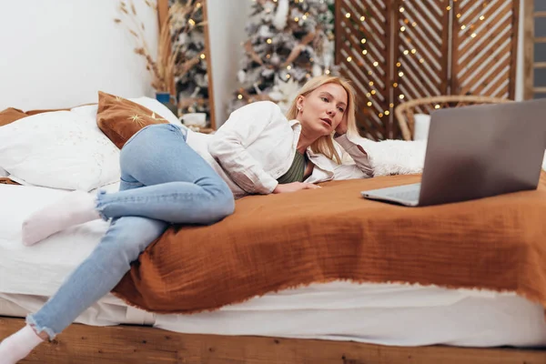 Νεαρή γυναίκα ξαπλωμένη στο κρεβάτι με φορητό υπολογιστή τα Χριστούγεννα — Φωτογραφία Αρχείου