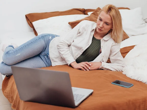Νεαρή γυναίκα ξαπλωμένη στο κρεβάτι της κοιτάζοντας την οθόνη του φορητού της υπολογιστή — Φωτογραφία Αρχείου