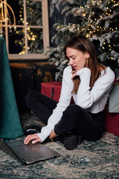 Νεαρή γυναίκα στο σπίτι δίπλα στο χριστουγεννιάτικο δέντρο κάθεται με φορητό υπολογιστή — Φωτογραφία Αρχείου