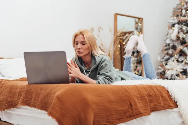 Genç bir kadın, Noel zamanı dizüstü bilgisayarına bakarak yatağa uzanıyor. — Stok fotoğraf