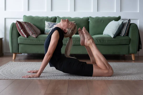Frau, die zu Hause trainiert, ihren Rücken wölbt und ihre Zehen nach ihrem Kopf streckt. — Stockfoto