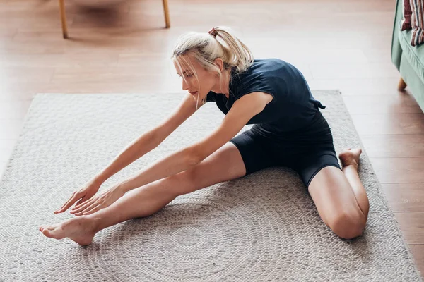 Frau übt zu Hause, macht Dehnübungen vorwärts, während sie auf dem Boden sitzt — Stockfoto