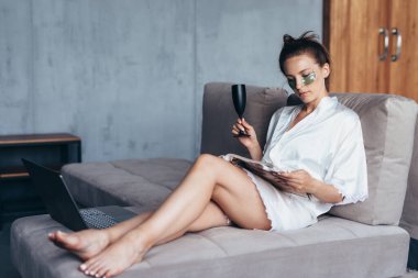 Kanepede oturan genç bir kadın. Gözlerinin altında yamalar ve dergiler var.