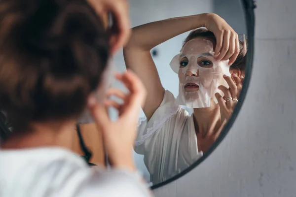 Woman applies sheet mask to her face in front of the mirror — Photo