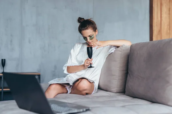 Woman with patches under her eyes sits on the couch and reads a magazine — 图库照片