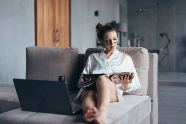 Woman with patches under her eyes sits on the couch and reads a magazine — Stock Photo, Image