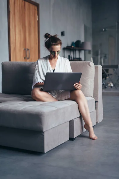 女人穿着睡衣，拿着笔记本电脑，坐在沙发上 — 图库照片