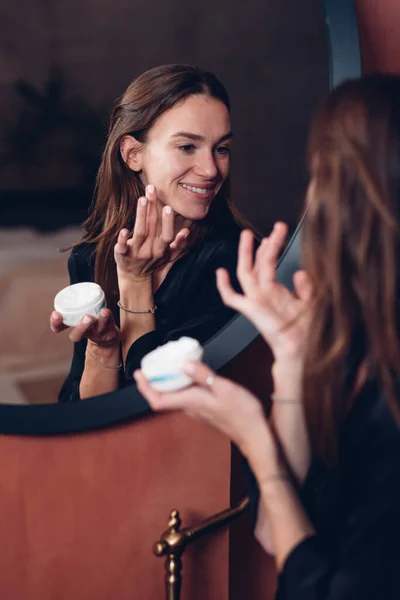 Young woman in front of the mirror putting cream on her face — Stock Photo, Image